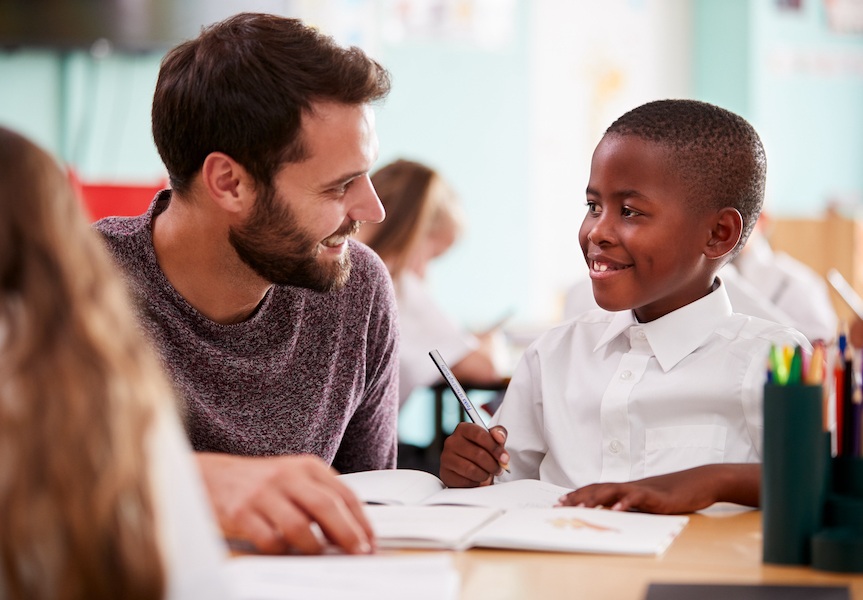 Elementary School Teacher Giving Male Pupil Wearing Uniform One To One Support In Classroom